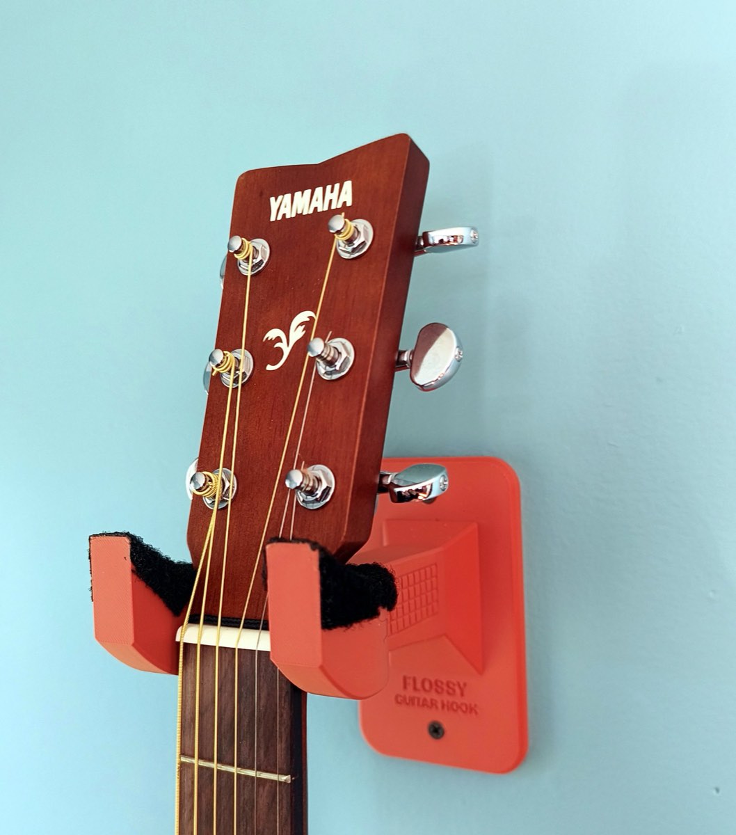 A printed Flossy Guitar Hook on a blue wall, holding an acoustic guitar by the neck just under the headstock